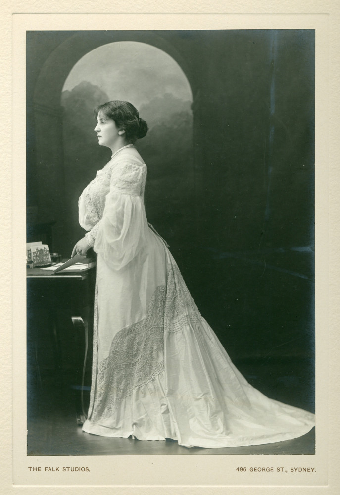 Photographie d'Hélène Paderewska en robe blanche, debout de profil devant une table, par The Falk Studios, Sydney, en 1904