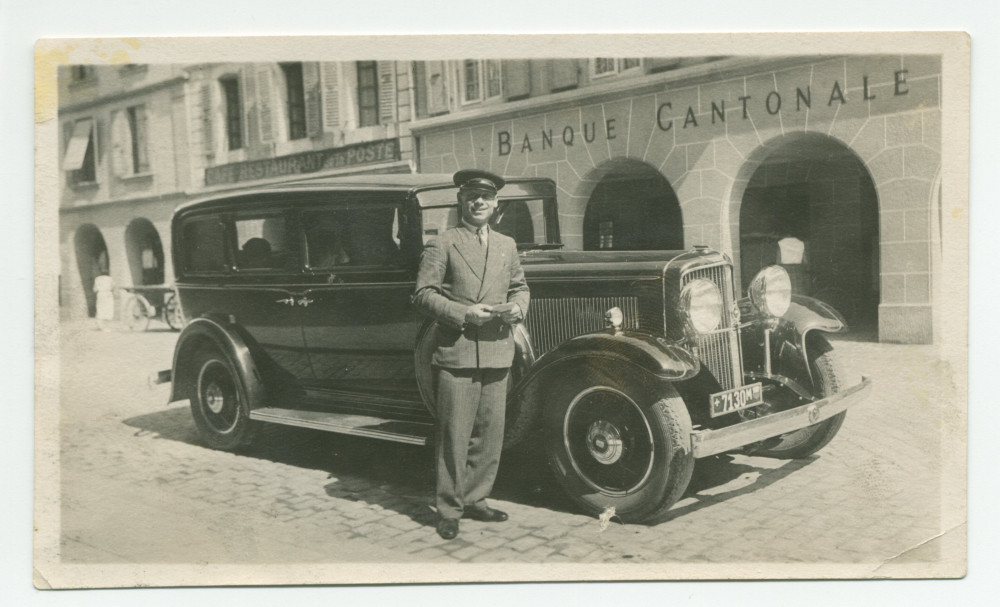 Photographie du chauffeur de Paderewski, Silvio Mongini, en costume et casquette, debout devant l'automobile du maître, sur la Grand-Rue de Morges, devant les bureaux de la Banque Cantonale Vaudoise