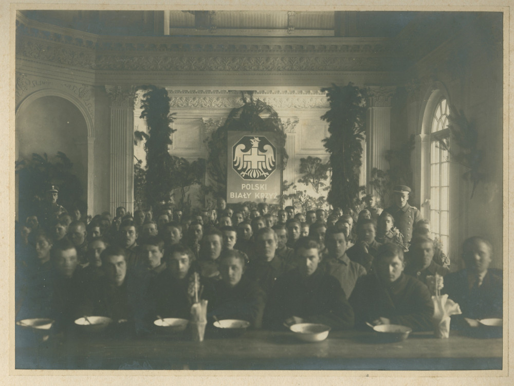 Photographie de soldats (ou prisonniers?) mangeant dans une cantine de la Croix Blanche polonaise (avec son insigne placardé en grand au fond), encadrés par deux militaires en uniforme, en 1918