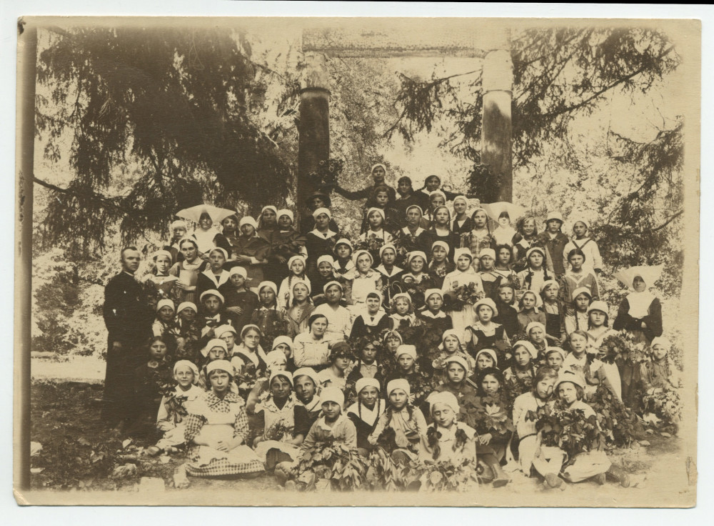 Photographie de groupe, sous des arbres, encadrées de religieux, des filles du Centre éducatif Sainte-Thérèse de Powisle, dans la banlieue de Varsovie (rue Lipowa 14), en 1921 – légende manuscrite au verso avec tampon de la Croix Blanche polonaise