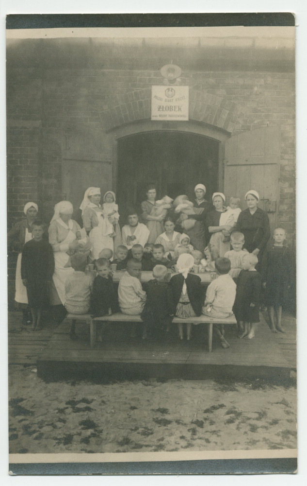 Photographie d'enfants assis à une table, encadré par des infirmières et des nurses, devant la porte d'entrée d'une crèche «Hélène Paderewska» de la Croix Blanche polonaise – tirage carte postale