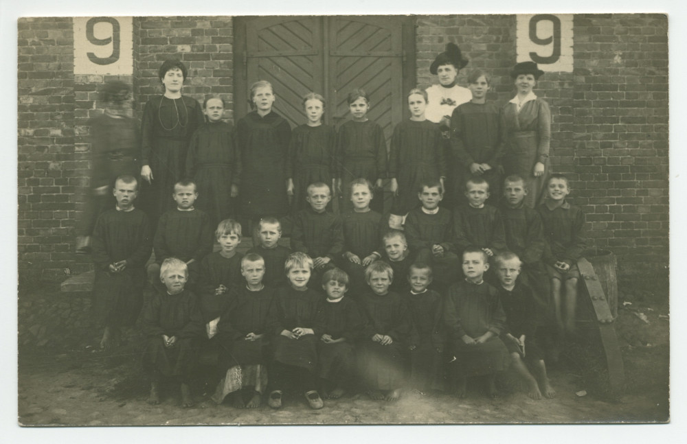 Photographie d'enfants en uniforme noir posant devant le mur en briques d'une crèche de la Croix Blanche polonaise, encadrés par quatre dames – tirage carte postale