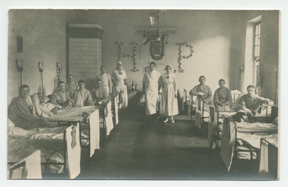Photographie d'hommes convalescents dans une chambre d'hôpital tenu par la Croix Blanche polonaise, avec sur le mur du fond des décorations en hommage à sa fondatrice Hélène Paderewska («HP») – tirage carte postale