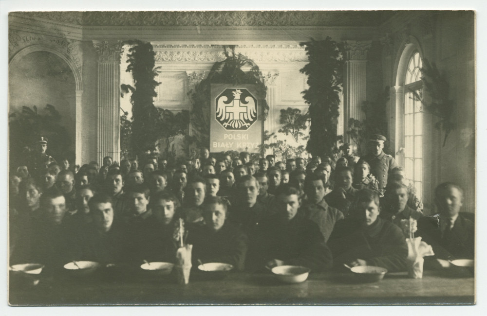 Photographie de jeunes hommes (soldats?) recevant à manger dans une grande salle décorée de branches de sapin (Noël), encadrés par deux hommes en uniforme, sous l'écusson imposant de la Croix Blanche polonaise – tirage carte postale