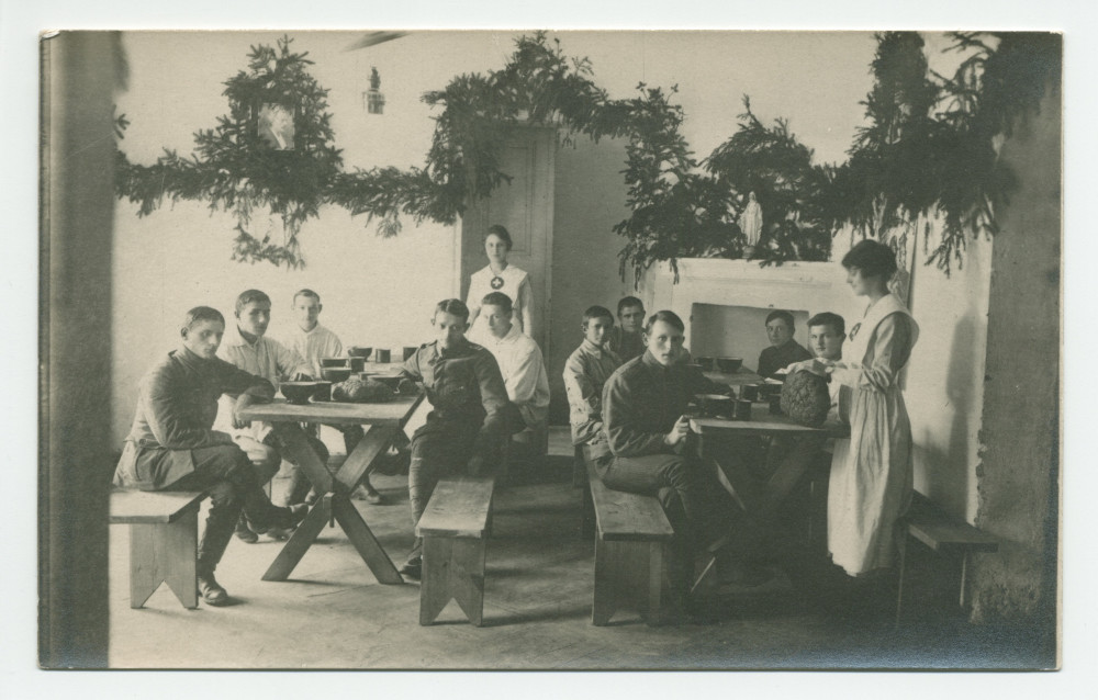 Photographie de jeunes soldats recevant à manger d'infirmières de la Croix Blanche polonaise dans un décor de branches de sapin (Noël?), avec au mur un portrait d'Ignace Paderewski – tirage carte postale