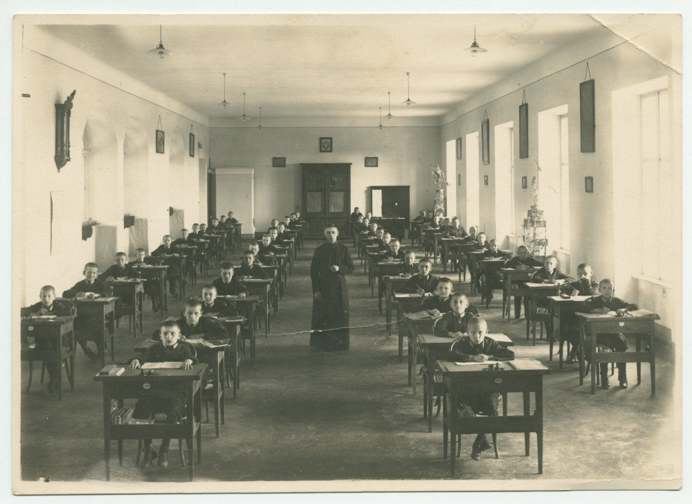 Photographie d'une salle de classe composée de jeunes garçons le crâne rasé, sous la supervision d'un ecclésiastique, dans une école patronnée sans doute par la Croix Blanche polonaise – tirage carte postale