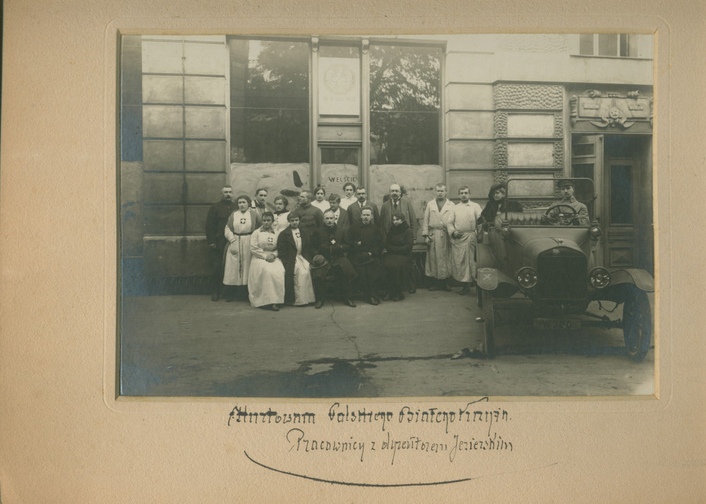 Photographie des employés de la Croix Blanche polonaise devant l'entrée des magasins de Varsovie (probablement situés dans le quartier de Wilanów), avec voiture et chauffeur (transportant vraisemblablement Mme Rembielin?ska