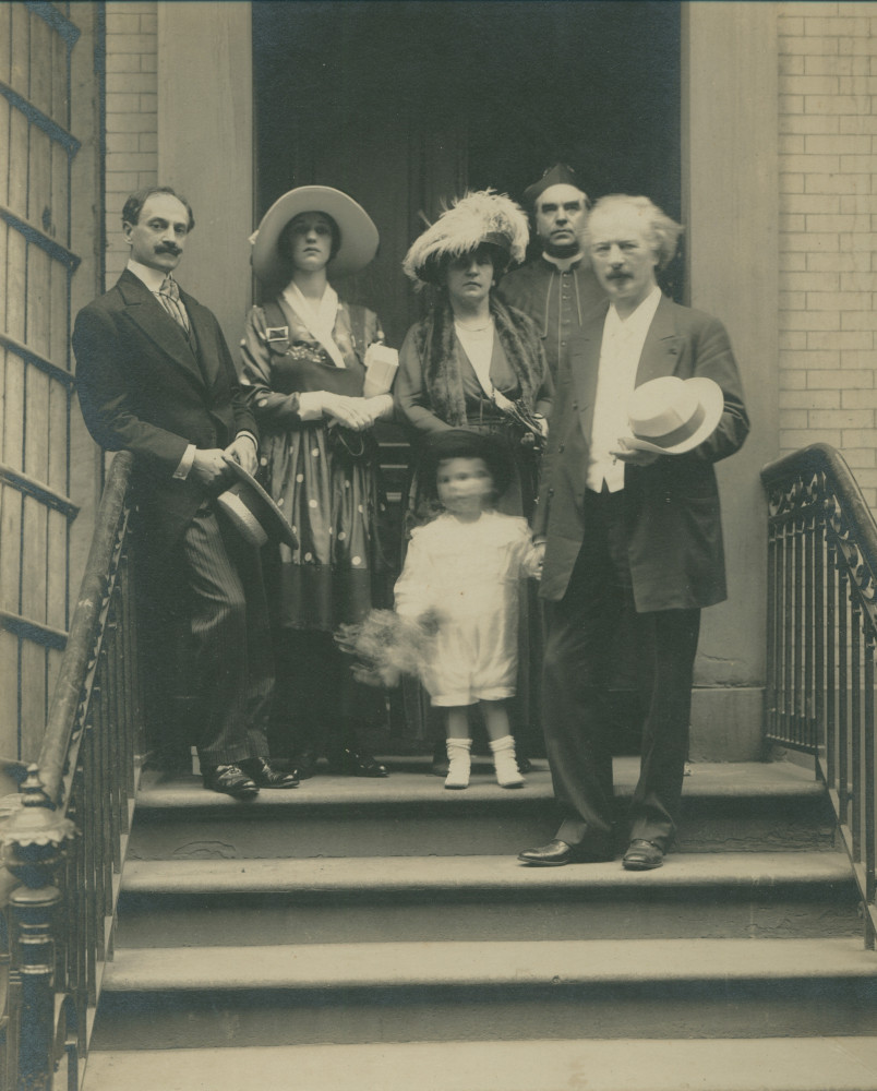 Photographie d'Hélène et Ignace Paderewski sur un perron à New York (?) avec un enfant (flou) tenant la main d'Ignace, un homme, une femme et un religieux en arrière-plan