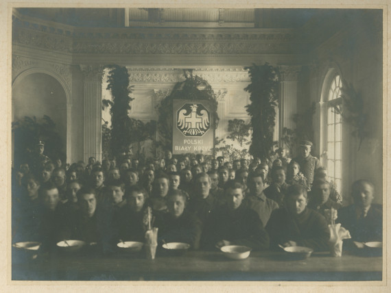 Photographie de soldats (ou prisonniers?) mangeant dans une cantine de la Croix Blanche polonaise (avec son insigne placardé en grand au fond), encadrés par deux militaires en uniforme, en 1918