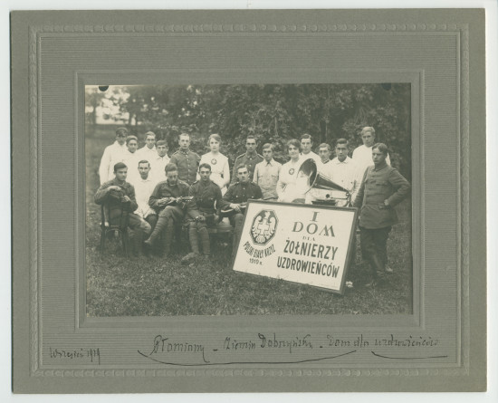 Photographie de soldats convalescents (dont deux jouant de la guitare) en compagnie d'infirmières dans le jardin de la maison de convalescence de la Croix Blanche polonaise à Plomiany, avec pancarte de l'établissement et gramophone, en septembre 1919