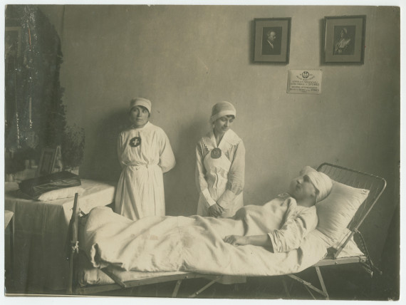 Photographie d'un soldat (?) blessé pris en charge par deux infirmières de la Croix Blanche polonaise, au sein de l'Hôpital «Hélène Paderewska», sous les portraits d'Ignace et d'Hélène (en costume de la Croix Blanche)