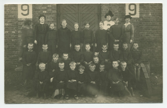 Photographie d'enfants en uniforme noir posant devant le mur en briques d'une crèche de la Croix Blanche polonaise, encadrés par quatre dames – tirage carte postale