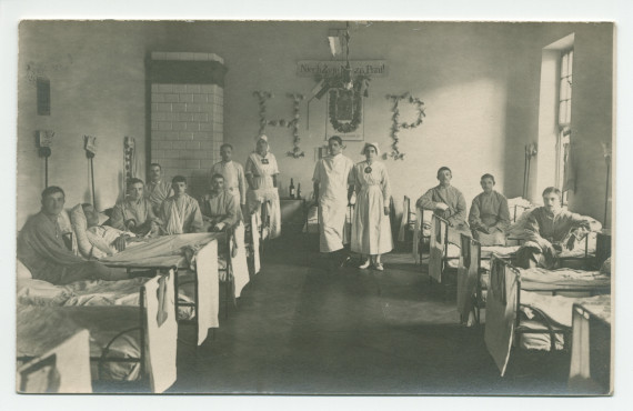 Photographie d'hommes convalescents dans une chambre d'hôpital tenu par la Croix Blanche polonaise, avec sur le mur du fond des décorations en hommage à sa fondatrice Hélène Paderewska («HP») – tirage carte postale