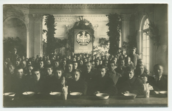 Photographie de jeunes hommes (soldats?) recevant à manger dans une grande salle décorée de branches de sapin (Noël), encadrés par deux hommes en uniforme, sous l'écusson imposant de la Croix Blanche polonaise – tirage carte postale