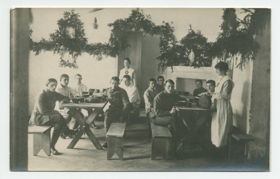 Photographie de jeunes soldats recevant à manger d'infirmières de la Croix Blanche polonaise dans un décor de branches de sapin (Noël?), avec au mur un portrait d'Ignace Paderewski – tirage carte postale