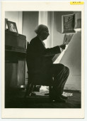 Carte postale de Paderewski lisant le journal – photographie de Lucien Aigner réalisée dans sa chambre de l'Hôtel Buckingham à New York au printemps 1941 (avec la photographie d'Ernest Schelling sur le piano droit)
