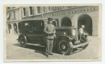 Photographie du chauffeur de Paderewski, Silvio Mongini, en costume et casquette, debout devant l'automobile du maître, sur la Grand-Rue de Morges, devant les bureaux de la Banque Cantonale Vaudoise