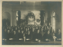 Photographie de soldats (ou prisonniers?) mangeant dans une cantine de la Croix Blanche polonaise (avec son insigne placardé en grand au fond), encadrés par deux militaires en uniforme, en 1918