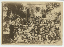 Photographie de groupe, sous des arbres, encadrées de religieux, des filles du Centre éducatif Sainte-Thérèse de Powisle, dans la banlieue de Varsovie (rue Lipowa 14), en 1921 – légende manuscrite au verso avec tampon de la Croix Blanche polonaise