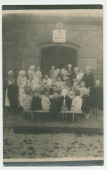 Photographie d'enfants assis à une table, encadré par des infirmières et des nurses, devant la porte d'entrée d'une crèche «Hélène Paderewska» de la Croix Blanche polonaise – tirage carte postale