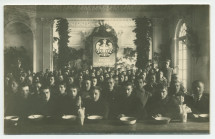 Photographie de jeunes hommes (soldats?) recevant à manger dans une grande salle décorée de branches de sapin (Noël), encadrés par deux hommes en uniforme, sous l'écusson imposant de la Croix Blanche polonaise – tirage carte postale
